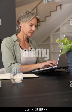 Woman typing on laptop at home workspace wearing light green overshirt near plant and notebook. Focus, productivity, calm, minimalist, modern, office, Stock Photo