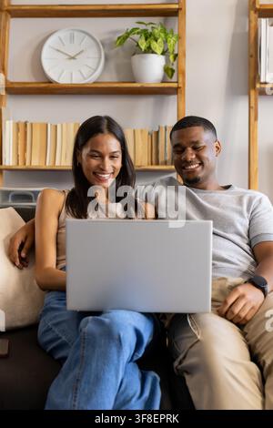 African American couple sitting on sofa at home using silver laptop with phone nearby, casual wear. Man, woman, livingroom, notebook, bookshelf, green Stock Photo