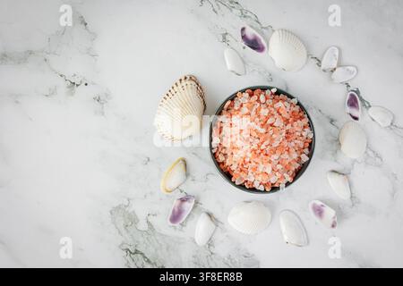 Coarse pink rock salt and assorted seashells arranged on a white marble studio background create an airy coastal wellness scene with calm mood and cop Stock Photo