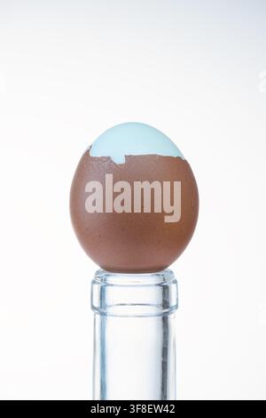 Brown egg with cracked top shell revealing white beneath, balanced on glass bottle Stock Photo