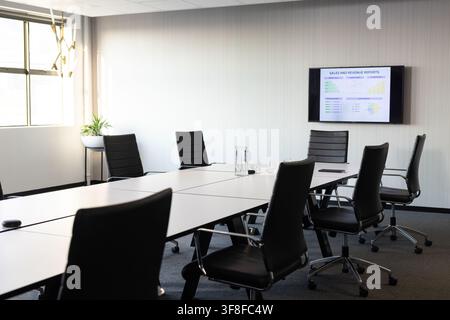 White conference table sitting in meeting room, black chairs circling, monitor showing charts Stock Photo