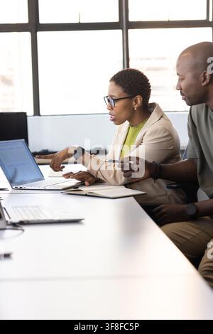 African American coworkers leaning forward collaborating on laptop at office table, woman in blazer Stock Photo