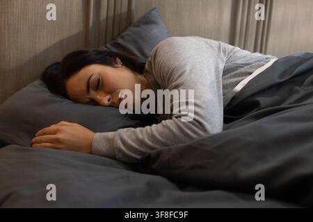 Woman lying sideways in bedroom wearing light gray top, resting head on pillow under dark duvet. Relaxation, sleep, cozy, minimalist, neutral, ambienc Stock Photo
