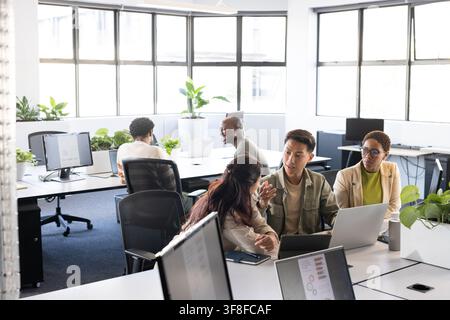Diverse coworkers collaborating around laptop and tablet in bright office, wearing business attire Stock Photo