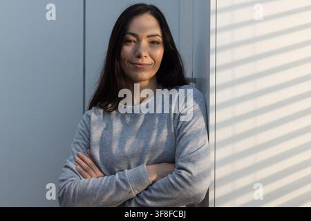 Woman leaning against light-colored panel near window, blinds casting striped shadows on sweatshirt Stock Photo