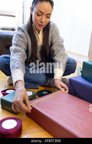 Asian woman wearing gray sweater, headband wrapping large red present on floor placing gold ribbon Stock Photo