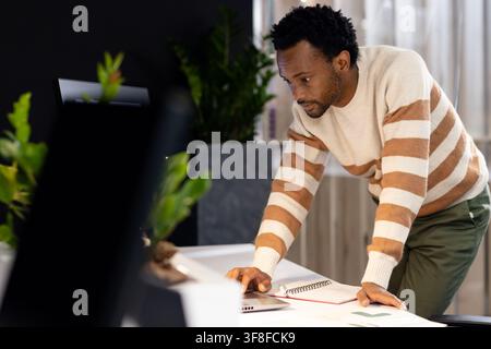 African American man leaning over white desk at office in cream sweater using laptop, notebook Stock Photo