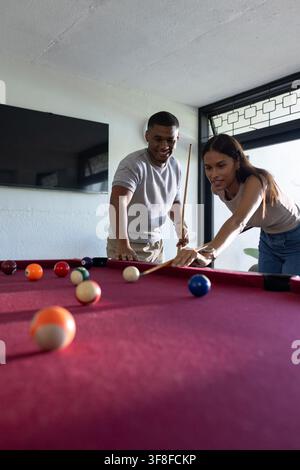 African American couple playing pool at home on red-felt table, aiming with cues and cue bridge. Billiards, gameroom, sunlit, casual, focus, residenti Stock Photo