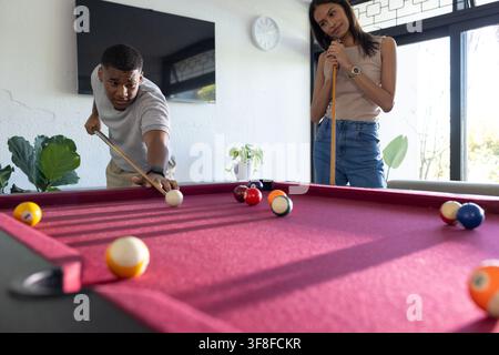 African couple playing billiards in home game room on burgundy pool table with cues. Pair, casual, leisure, sunlit, interior, windows, contemporary Stock Photo