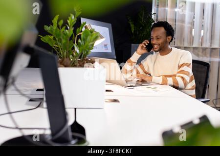African American man in striped sweater speaking on phone, typing laptop at open office, copy space. Professional, desk, plants, chart, monitor, works Stock Photo