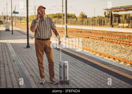 Happy senior man is standing at railway station and talking on mobile phone. Senior people traveling. Stock Photo