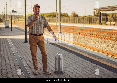 Happy senior man is standing at railway station and talking on smartphone. Senior people traveling. Stock Photo