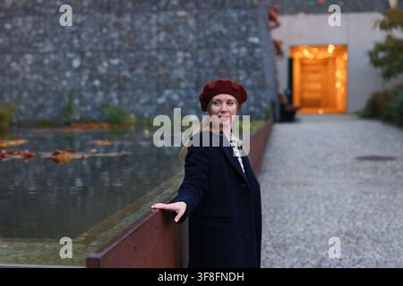 Young stylish woman in red beret and dark coat posing outdoors by a reflecting pool in modern urban architecture. Stock Photo