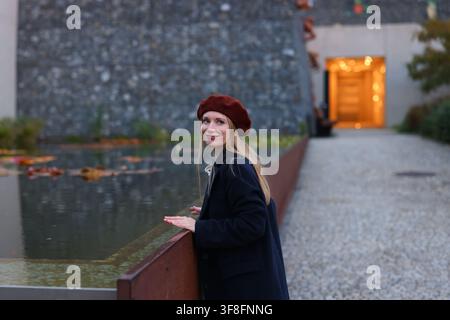 Fashionable young woman wearing red beret and black coat standing near water in an urban setting during autumn. Stock Photo