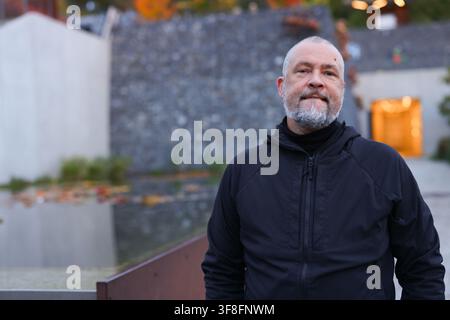 Mature bearded man in black jacket standing outdoors in a modern urban setting, confident male lifestyle portrait. Stock Photo