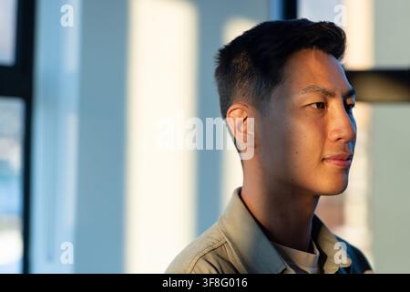Asian mid-adult man standing near tall windows inside bright room in beige jacket, copy space Stock Photo
