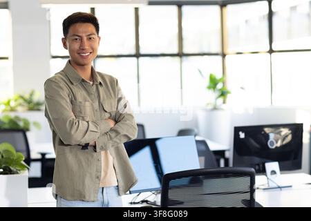 Asian man standing arms crossed smiling in office wearing olive tee computer monitors visible. Workspace, modern, screens, desks, greenery, bright, mi Stock Photo