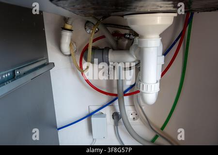 Complex network of plastic drain pipes flexible hoses and colored water supply lines installed under domestic sink with power outlets visible. Stock Photo