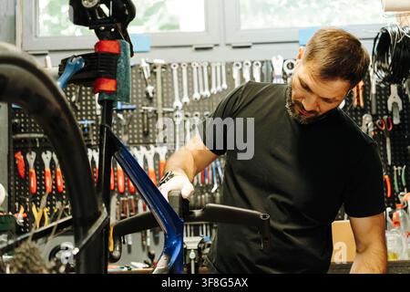 Male bicycle mechanic performs maintenance on a road bike mounted in a workshop stand. Professional tools are arranged on the wall behind the worker. Stock Photo
