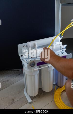 Man hand inserting yellow plastic hose into reverse osmosis water filter system during domestic installation process on wooden floor background. Stock Photo