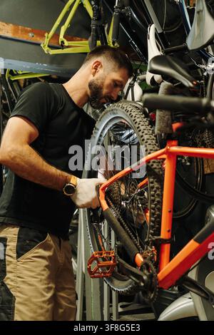 Male bicycle mechanic works on an orange mountain bike surrounded by bicycles and parts. Professional maintenance focuses on the rear wheel and Stock Photo