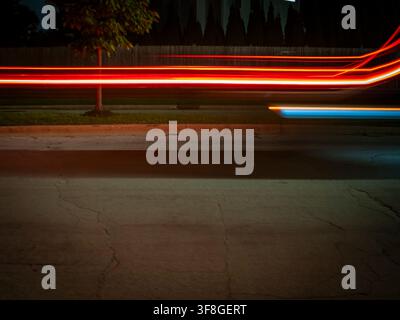 Long exposure night photography showing vibrant red and blue light trails from moving vehicles speeding past a suburban street and trees Stock Photo