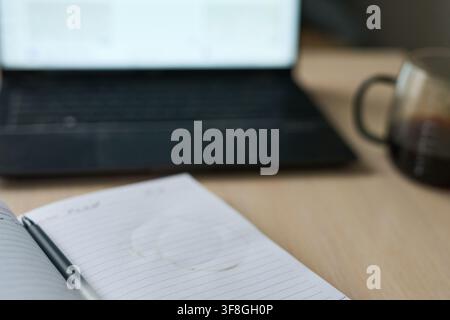 Closeup Lined Notebook Coffee Ring And Pen With Blurred Laptop Keyboard And Glass Mug On Wooden Desk In Home Office Workspace Under Soft Daylight Showing Subtle Surface Texture. Stock Photo