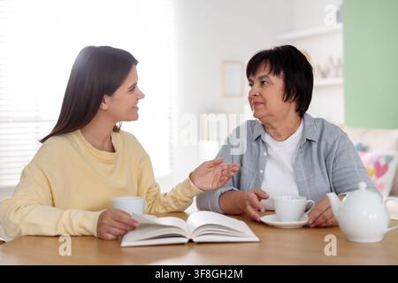 Caregiver and senior woman enjoying tea while reading book at wooden table indoors Stock Photo