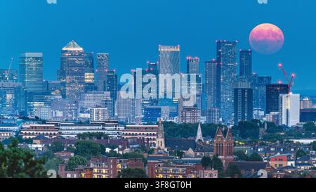 View of the London skyline at dusk featuring the Canary Wharf skyscrapers illuminated against a deep blue sky with a large full moon rising over the c Stock Photo