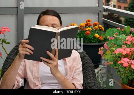 Middle aged woman sitting in wicker chair reading hardcover book with potted flowers in background on balcony. Stock Photo