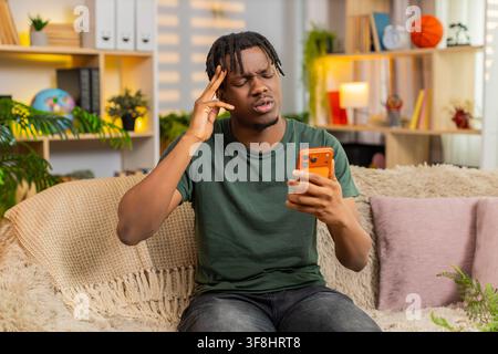 African American young man on home sofa looking at smartphone with sad face after bad news and online game loss. Black guy on home sofa feels upset while reading screen and reacting to online defeat. Stock Photo
