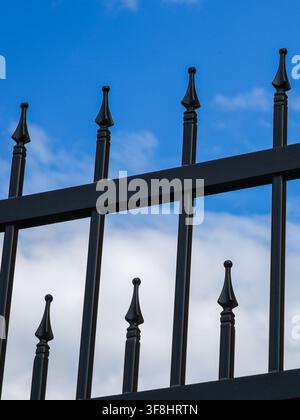 Close-up view of black metal fence with decorative spearhead pickets silhouetted against a vivid blue sky with soft white clouds in daylight Stock Photo