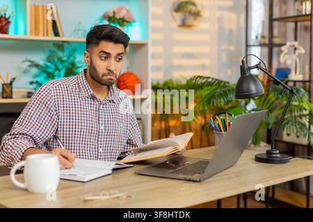 Indian man on home table reads book writes notes and checks laptop screen during focused online study in living room. Arabian guy on home table studies with calm focus in daily home learning session. Stock Photo
