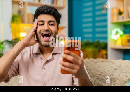 Indian man on home sofa watches smartphone with wide eyes open mouth wow reaction after unexpected news. Arabian guy on home sofa laughs with shocked Stock Photo