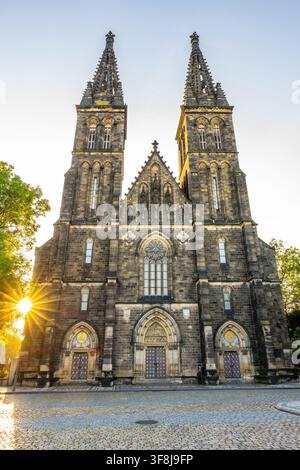 The Basilica of St. Peter and Paul stands tall in Vysehrad. Bright sunlight shines from behind the church, highlighting its towers and stone details. The area is quiet and open. Stock Photo