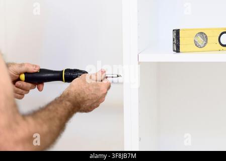 Assembling flat-pack white wardrobe furniture in a bedroom. Close-up of male hands using a screwdriver and bubble level to install shelving and align Stock Photo