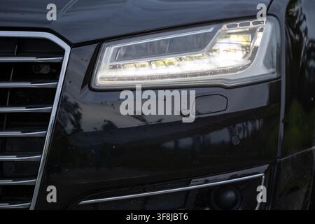 Close-up view of a luxury black car front showing the illuminated LED headlight, chrome grille and glossy bumper details, conveying modern automotive Stock Photo