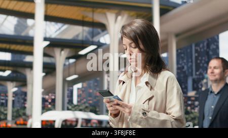 Traveler walking on street looks down at phone screen and follows updates for upcoming bus connection. Woman navigating coach transit hub using smartphone to confirm directions and departure details Stock Photo