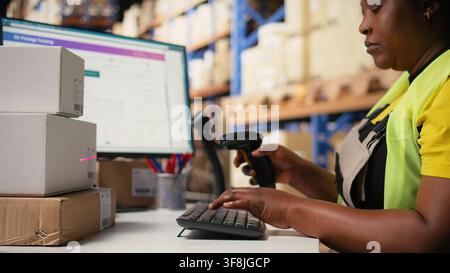 Female worker managing barcodes and shipment details scanning tasks for online shopping orders, goods with shipping labels awb numbers on boxes. Distribution hub staff in hi vis vest. Camera A. Stock Photo