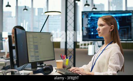 Female technician coding with AI language model assistance in startup office, using PC. Intern using computer, integrating artificial intelligence LLM assistant suggestions into code editor, camera A Stock Photo