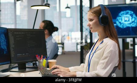 Young female programmer in office reviewing AI system metrics, listening music, monitoring performance dashboards. Junior dev wearing headphones tuning neural networks using tools on PC, camera A Stock Photo