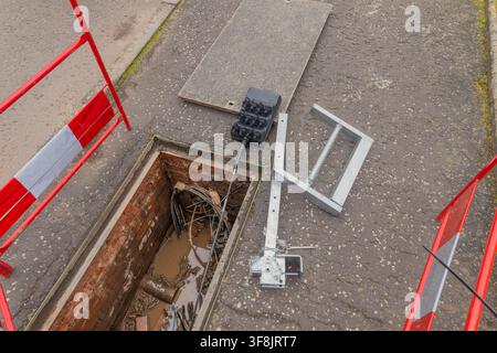 Open brick-lined telecoms chamber in the road reveals cabling and ducting in muddy water, with a black multi-duct cable seal resting on the tarmac bes Stock Photo