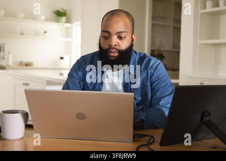 Bearded man sitting at wooden table in bright kitchen, denim shirt, working on laptop with tablet Stock Photo