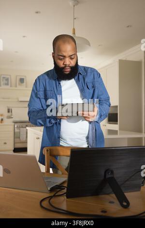 African adult man standing at table in kitchen, using tablet and laptop, wearing denim overshirt Stock Photo