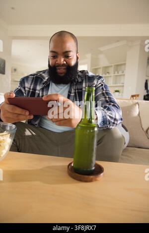 Adult African American man sitting on couch at home holding phone, wearing plaid shirt, olive pants Stock Photo
