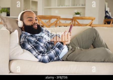Man reclining on sofa at home, in plaid shirt, holding smartphone, listening on white headphones Stock Photo
