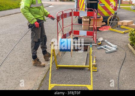 Worker in a yellow-green high-visibility jacket and red gloves feeds black fibre optic cable from a blue rope drum mounted on a yellow cable jack. A r Stock Photo