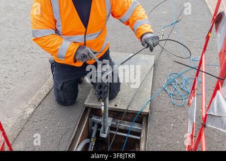 Engineer in an orange high-visibility jacket kneels at an open telecoms chamber, feeding a cable rod through a black duct seal mounted on a galvanised Stock Photo
