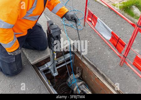 Telecoms worker in an orange high-visibility jacket handles blue draw rope at a black duct seal mounted on a galvanised stand over an open telecoms ch Stock Photo