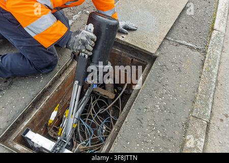 Telecoms worker in orange high-visibility jacket kneels and handles a large black cable seal head mounted on a galvanised stand over an open telecoms Stock Photo